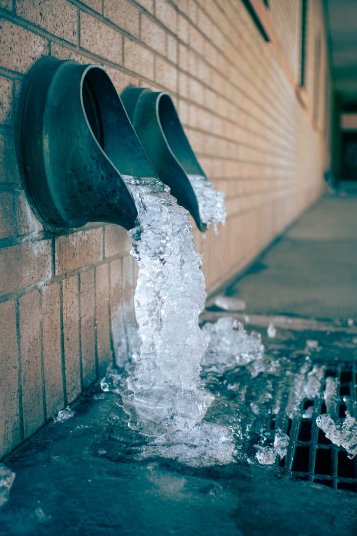 Close-up of frozen water flowing from building drain pipes into a grate.